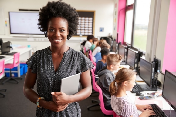 Portrait Of Female Teacher Holding Digital Tablet Teaching Line Of High School Students Sitting By Screens In Computer Class